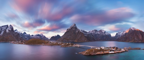Winter landscape with houses in village, snowy mountains, sea, blue cloudy sky reflected in water at sunrise. Beautiful Hamnoy and Reine fisherman village, Lofoten islands, Norway. Norwegian fjords