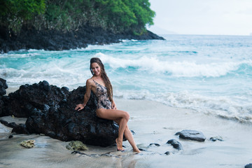 beautiful long-haired tanned girl of European appearance posing for the camera on the background of the ocean beach, stones and trees. Indonesia, Bali