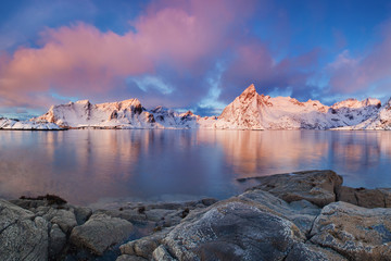 Winter landscape with houses in village, snowy mountains, sea, blue cloudy sky reflected in water at sunrise. Beautiful Hamnoy and Reine fisherman village, Lofoten islands, Norway. Norwegian fjords