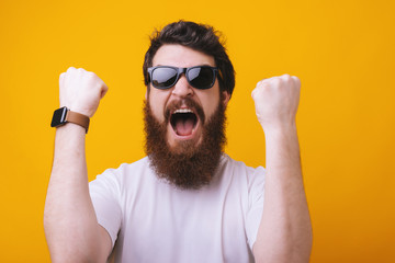 A handsome excited bearded man in sunglasses standing with fists up on yellow background