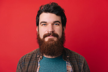 Shot of young handsome male with beard, has serious expression as listens to interlocutor, poses in studio against redbackground