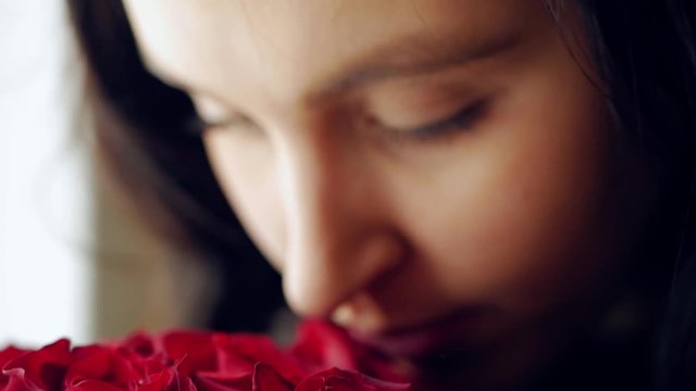 Beautiful girl sniffs a bouquet of red roses and then looks out the window. Closeup