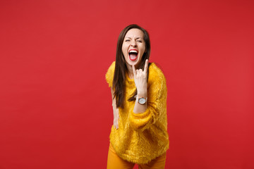 Screaming young woman in yellow fur sweater with horns up gesture, depicting heavy metal rock sign isolated on bright red background. People sincere emotions, lifestyle concept. Mock up copy space.