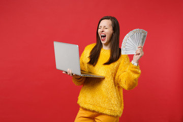 Happy young girl with closed eyes screaming holding laptop pc computer, fan of money in dollar banknotes, cash money isolated on red background. People emotions, lifestyle concept. Mock up copy space.