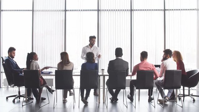 Achieving best results after annual refresher course. Indian young business mentor conducting a business training while standing in front of people sitting in row at meeting room.
