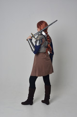 full length portrait of a  red haired girl wearing medieval warrior costume and steel armour, standing pose facing away from the camera on grey studio background.