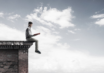 Man on roof edge reading book and cloudscape at background