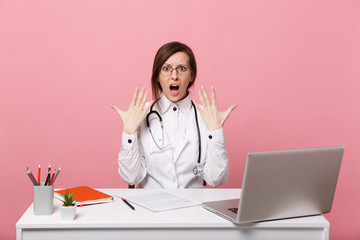 Tired mad female doctor sits at desk work on computer with medical document in hospital isolated on pastel pink wall background. Woman in medical gown glasses stethoscope. Healthcare medicine concept.