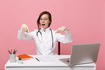 Beautiful female doctor sits at desk works on computer with medical document in hospital isolated on pastel pink wall background. Woman in medical gown glasses stethoscope. Healthcare medicine concept