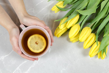 Female hands holding cup of tea with lemon slice and yellow tulips on a marble background. Flu risk in spring.