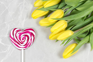 Romantic flat lay - swirly lollipop in a shape of a heart and yellow tulips on a marble background. Festive flat lay for Valentine's day, mother's day, 8 march.