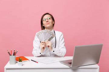 Female doctor sits at desk work on computer with medical document cash money in hospital isolated on pastel pink wall background. Woman in medical gown glasses stethoscope. Healthcare medicine concept