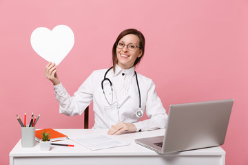 Female doctor sit at desk work on computer with medical document hold heart in hospital isolated on pastel pink wall background. Woman in medical gown glasses stethoscope. Healthcare medicine concept.