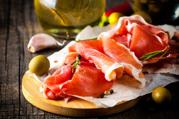 Closeup of thin slices of prosciutto with mixed olives and paprika on wooden cutting board