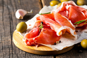 Closeup of thin slices of prosciutto with mixed olives and paprika on wooden cutting board