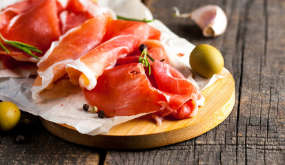 Closeup of thin slices of prosciutto with mixed olives and paprika on wooden cutting board