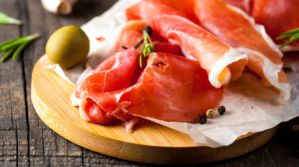 Closeup of thin slices of prosciutto with mixed olives and paprika on wooden cutting board