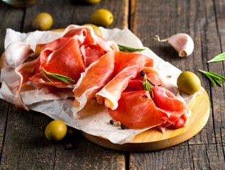 Closeup of thin slices of prosciutto with mixed olives and paprika on wooden cutting board