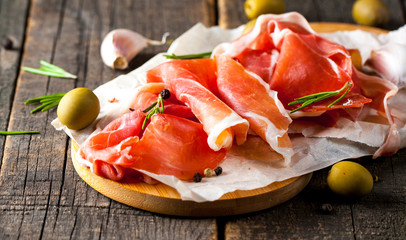 Closeup of thin slices of prosciutto with mixed olives and paprika on wooden cutting board
