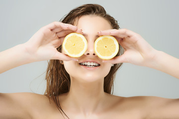 Creative portrait of a beautiful young women holding juicy lemon slices in front of eyes оn gray...