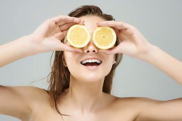 Creative portrait of a beautiful young women holding juicy lemon slices in front of eyes оn gray...
