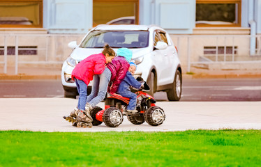 City Park, a funny boy rides on a toy electric car and his two older sisters riding on the rollers behind him on a warm autumn day. Having fun time with the family concept