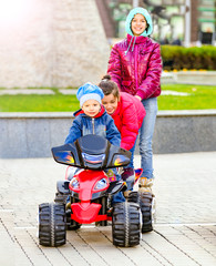 City Park, a funny boy rides on a toy electric car and his two older sisters riding on the rollers behind him on a warm autumn day. Having fun time with the family concept