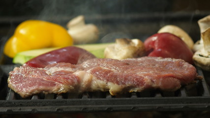 Steak of beef, pork or veal lies on the grill, in the background smoke from the grill and grilled vegetables, background is washed, vegetables on the grill, pepper, zucchini, mushrooms