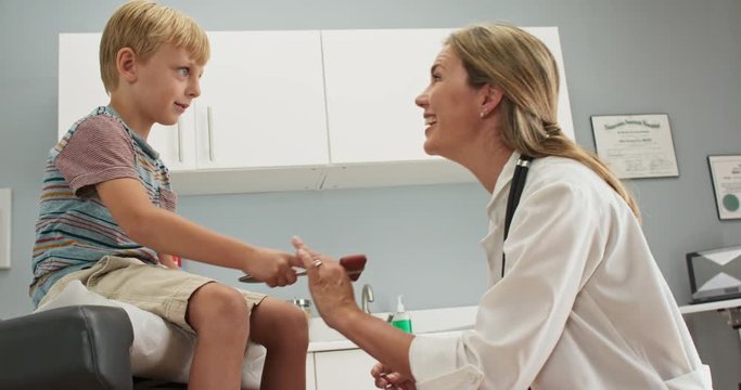 Little boy at pediatric office checking his reflexes with hammer while doctor watches. Female pediatrician showing patient how reflex hammer works. Slow motion 4k