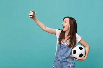 Surprised young girl football fan with soccer ball doing selfie shot on mobile phone isolated on blue turquoise background. People emotions, sport family leisure lifestyle concept. Mock up copy space.