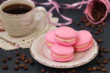 Pink macaroons are located on a plate on a dark background. In the background is a cup of coffee.