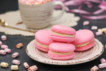 Pink macaroons are located on a plate on a dark background. In the background is a cup of coffee.