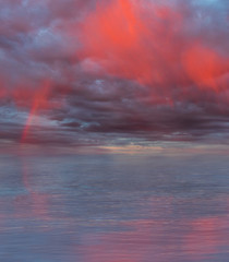 Fototapeta premium Dramatic cloudy sky with rainbow and red rain curtains reflecting on sea