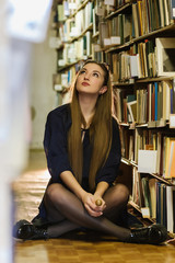 Beautiful young girl sitting among bookshelves and books on the floor in the library