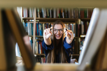 Beautiful young woman wearing glasses in the library among the bookshelves holding glasses and laughing, reading, studying.