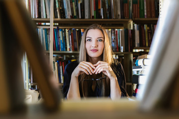 Beautiful young woman wearing glasses in the library among the bookshelves holding glasses and...