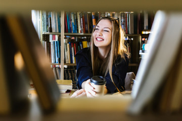 Beautiful young woman wearing glasses in the library among the bookshelves holding glasses and...