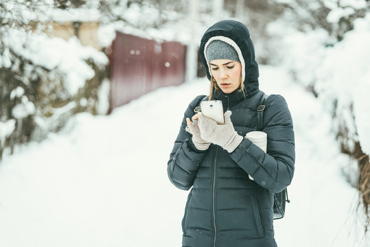 Beautiful woman dressed in a black winter jacket with mobile phone and thermos flask under her arm. Snow landscape background behind