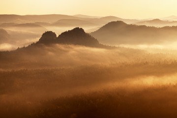 Sunrise in a beautiful mountain in Czech/Saxon Switzerland. Sandstone peaks increased from foggy background, the fog is orange to sun rays. Autumn or summer sunrise above misty and rocky valley