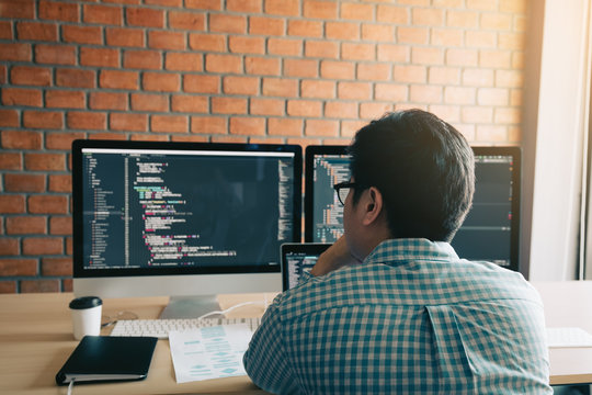 Rear view of contemporary website developer writing code for program and looking monitor screen with analyzing code.