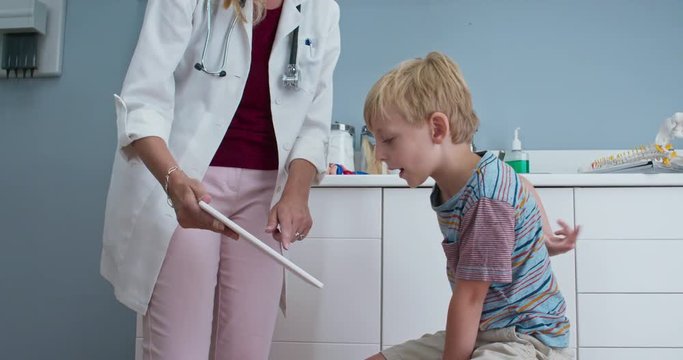 Female pediatrician showing tablet computer screen to child patient in exam room. Doctor explaining condition to little boy in exam room. Slow motion 4k