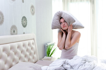 Sad unhappy woman suffering and disturbed from noisy neighbors and covering her ears with pillow while trying to falling sleep in bed at home in early morning.