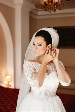 Beautiful and fashionable young brunette woman in the classic stylish wedding dress with a bridal veil on her head adjusting her earring and posing at interior