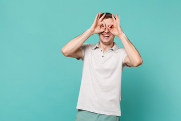 Laughing young man in casual clothes holding hands near eyes imitating glasses or binoculars isolated on blue turquoise wall background. People sincere emotions, lifestyle concept. Mock up copy space.
