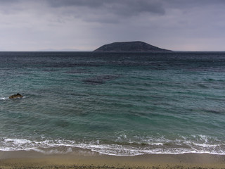 Small island, sea, clouded sky, and sand. Turquoise colored water and small wave foam on sandy beach with small island on background.