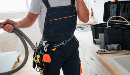 Cut view of man standing in kitchen and holding hose. He wear working uniform. Tools on belt. Guy...