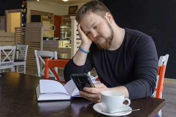 Handsome bearded man wearing black shirt looking at the screen of a mobile phone. A photo of a young handsome man freelancer sitting at the table with a mobile phone. Copy space