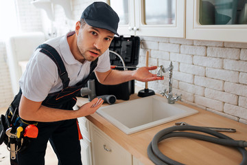 Amazed young man look on caerma and point on sink in kitchen. Worker has hose and toolbox on desk....