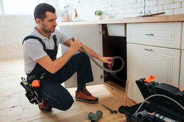 Concentrated young worker doing repairing in kitchen. He sit in squad pose and hold hose. Piece of...