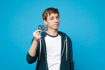 Portrait of handsome young man in casual clothes holding credit bank card isolated on blue wall background in studio. People sincere emotions, lifestyle concept. Mock up copy space. Advertising area.